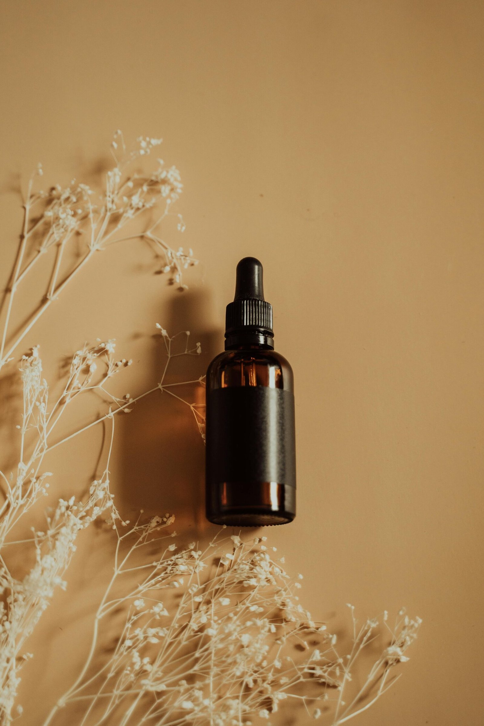 A minimalist flat lay of a brown dropper bottle beside delicate dried flowers on a beige background.